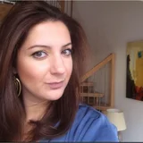 Portrait of smiling woman with wavy brown hair, hazel eyes, gold hoop earrings, blue top; wooden stairs, abstract art in background.